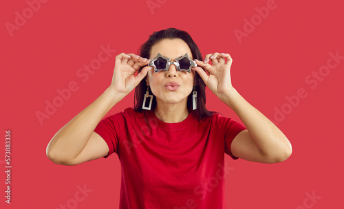 Cool and positive woman in star shape glasses makes an air kiss on vivid red background. Portrait of young woman who uses festival accessories and purses her lips for kiss while looking at camera.