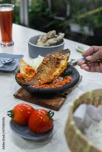 Close up sambal gami gurame or carp fish with sambal cooked in earthenware plate against wooden background. Served on wooden table with chili, spoon and fork as decoration