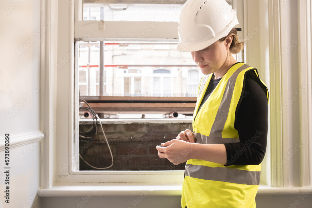 Chartered civil engineer lady using a calculator in front of a window in a construction site, wears yellow high visibility vest, ppe, personal protective equipment, site supervision, indoors Chartered civil engineer lady using a calculator in front of a window in a construction site, wears yellow high visibility vest, ppe, personal protective equipment, site supervision, indoors