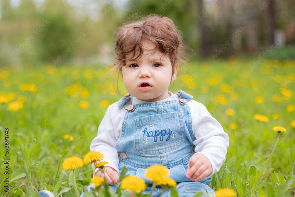 Happy little baby girl sitting on a green meadow with yellow flowers dandelions on the nature in the park Happy little baby girl sitting on a green meadow with yellow flowers dandelions on the nature in the park