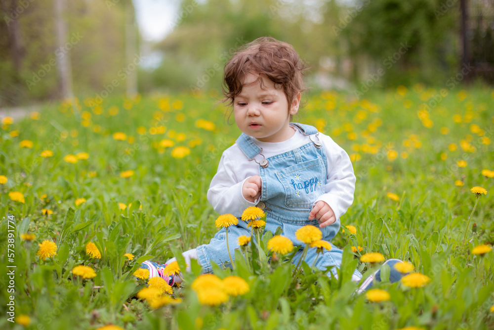 Happy little baby girl sitting on a green meadow with yellow flowers dandelions on the nature in the park Happy little baby girl sitting on a green meadow with yellow flowers dandelions on the nature in the park