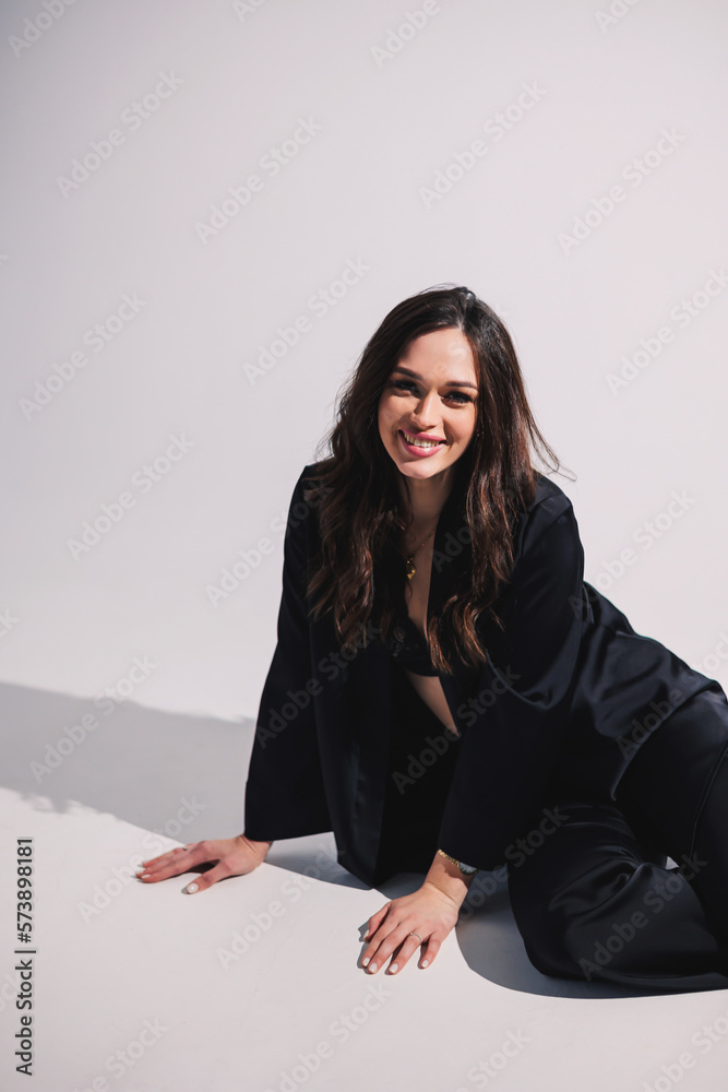 A stylish pretty young woman in a black classic suit is sitting on a white background. A beautiful woman in a jacket and make-up on her face