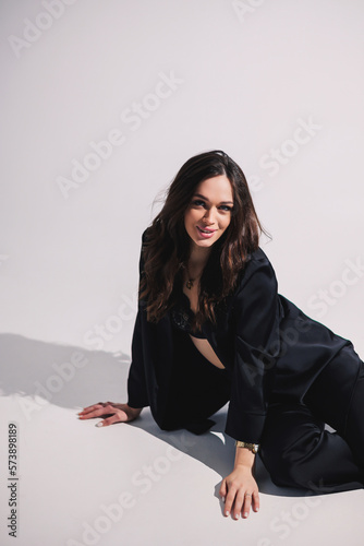 A stylish pretty young woman in a black classic suit is sitting on a white background. A beautiful woman in a jacket and make-up on her face