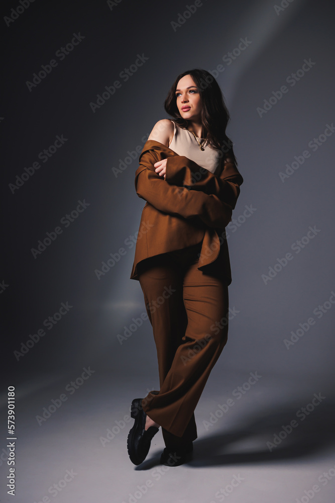 Stylish pretty young woman in brown classic suit sitting on gray background. Beautiful woman in a jacket and with makeup on her face