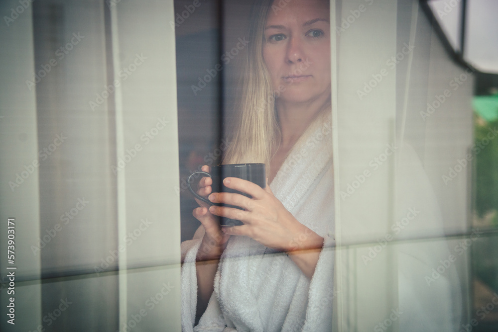 Woman in elegant robe drinking coffee in hotel room and standing near window. window reflection