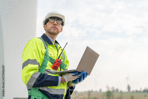 Male engineer in uniform with helmet safety using laptop inspection and maintenance of wind turbines in wind farms to generate renewable energy.