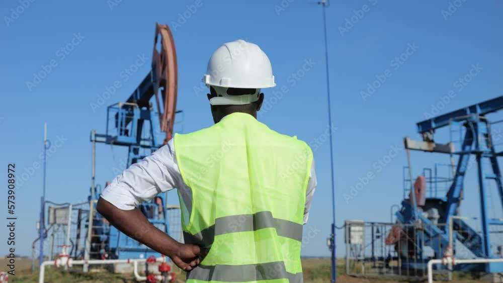 African American man controls beam-balanced pumping unit operation on sunny meadow. Black developer wearing working clothes explores production of oil African American man controls beam-balanced pumping unit operation on sunny meadow. Black developer wearing working clothes explores production of oil