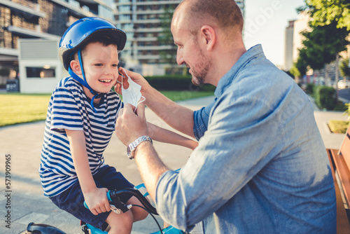 An adorable smiling little boy sitting on a bicycle, looking at his dad who is putting a protective face mask on his face.