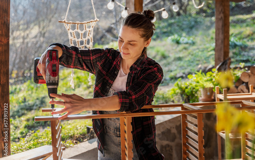 Obraz na płótnie Young woman in a plaid shirt and denim overalls assembling wooden bamboo furniture for a terrace in a country house, diy concept w salonie