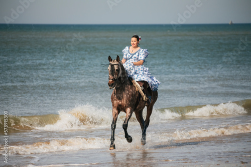 Woman in flamenco dress riding a horse galloping along the shore of the beach on a sunny day