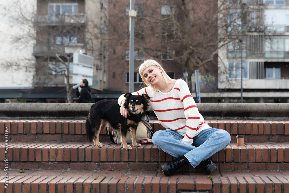 Obraz premium Young beautiful hipster woman is sitting on the stairs in the street with her foster dog she adopted from an animal shelter giving him love and attention