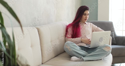 Young woman with colored hair is using laptop computer, sitting on the white sofa in the cozy living room. She is watching news on laptop and working or studing at home workplace. Home office concept. photo
