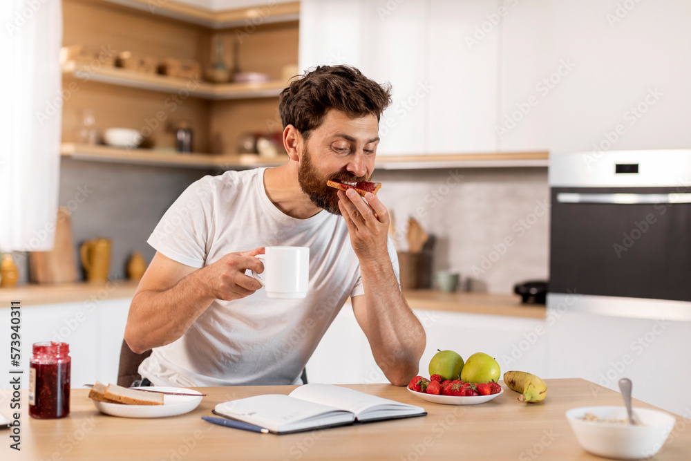 Busy handsome adult caucasian male in white t-shirt eats sandwich with jam, drinks coffee, reads book Busy handsome adult caucasian male in white t-shirt eats sandwich with jam, drinks coffee, reads book