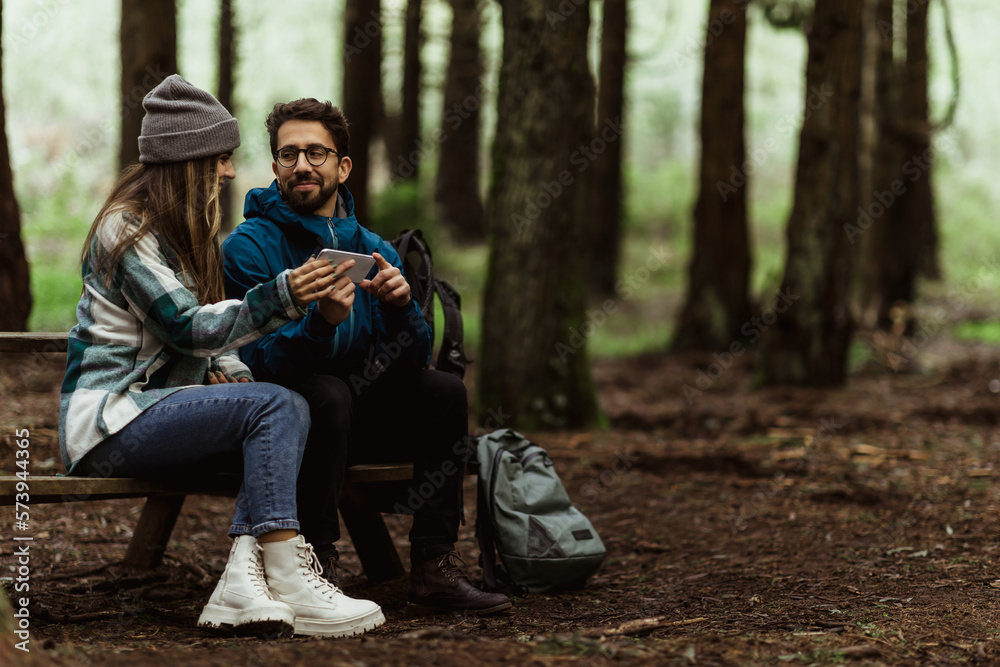 Smiling millennial caucasian guy in jacket show smartphone to wife, sit on bench, relaxing in cold forest Smiling millennial caucasian guy in jacket show smartphone to wife, sit on bench, relaxing in cold forest