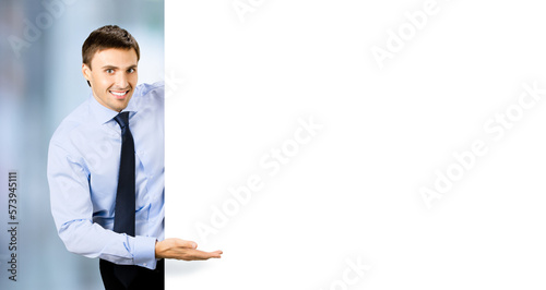 Portrait picture of young business man bank manager in confident cloth, necktie stand behind show empty white banner signboard billboard with copy space text area. Blurred office background