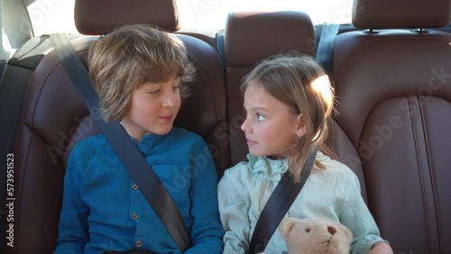 Two little cute children sitting in back seat of car while traveling using tablet smiling and talking. Beautiful blonde boy and lovely pretty girl fastened with safety belts. Car trip concept.
