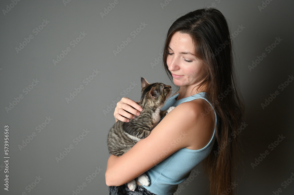 Woman hugging her kitten. Striped kitten lies on on the shoulder of a woman on gray background. Woman hugging her kitten. Striped kitten lies on on the shoulder of a woman on gray background.