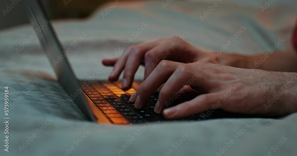 A man is typing on a laptop keyboard while lying on a bed in the evening, close-up of male hands, no face, unrecognizable identity