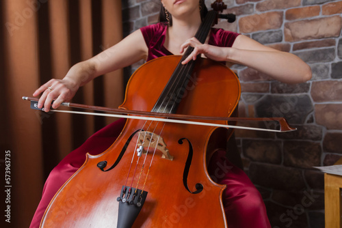 A creative cellist in red dress is playing the cello against a brick wall background.
