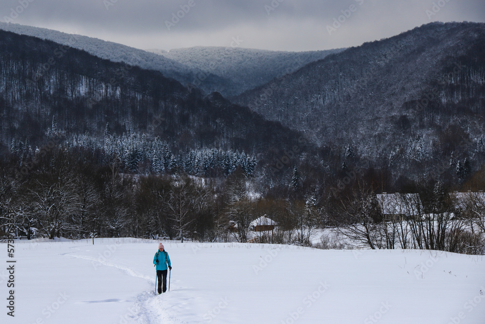 Hiker girl walks along snowy field with mighty mountains in background; hiking in cold winter weather Hiker girl walks along snowy field with mighty mountains in background; hiking in cold winter weather