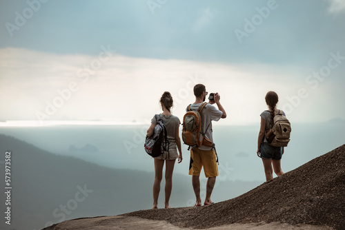 Print op canvas Three young tourists with backpacks stands on mountain top and looks at sea view