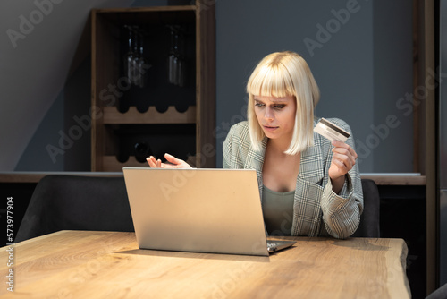 Young shocked frustrated woman holding bank credit card annoyed that she has reached her account limit and has no more money, she is refused online shopping and ordering on laptop computer