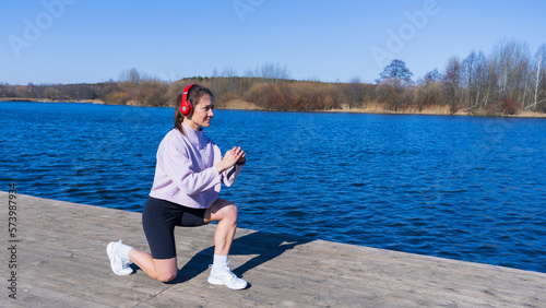 A young woman in a tracksuit listens to music and trains outdoors.A pond with clear water on the background. Fresh air in the park © makedonski2015