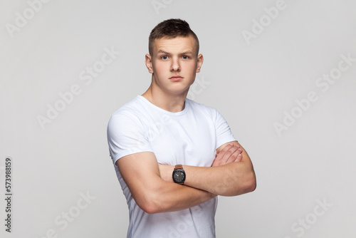 Serious teenager boy standing with folded hands, looking at camera with strict expression.