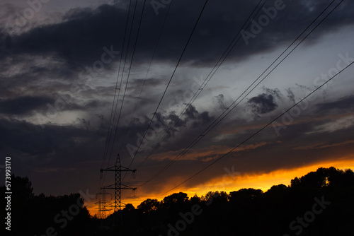 silhouettes of electricity pylons with high voltage cables in the middle of a forest at sunset