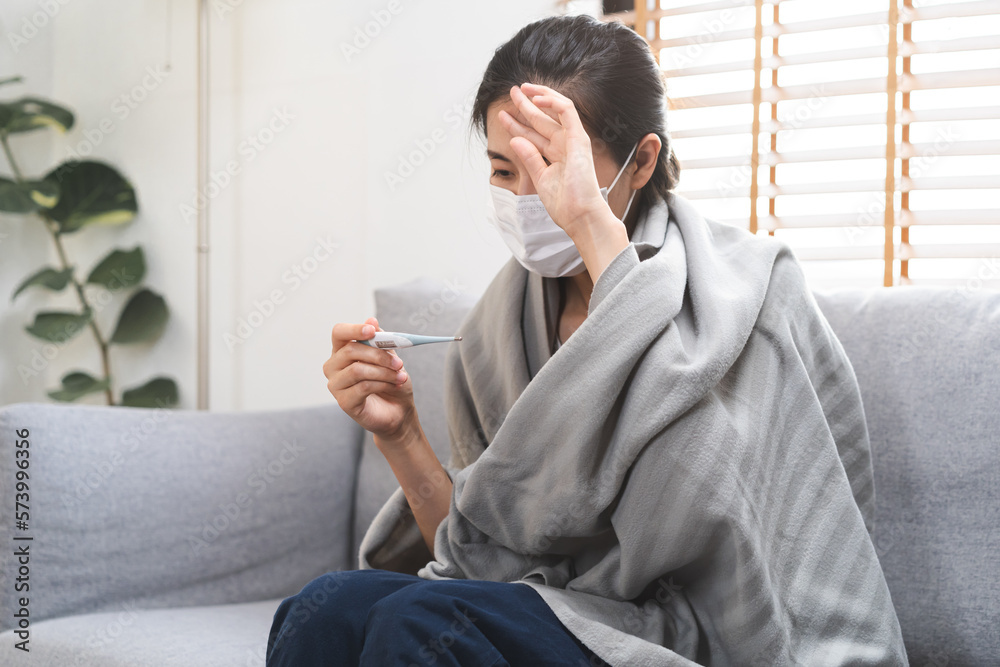 Sick, influenza asian young woman, girl wearing face mask in headache have a fever, flu and check thermometer measure body temperature, feel illness sitting on sofa bed at home. Health care, covid-19.