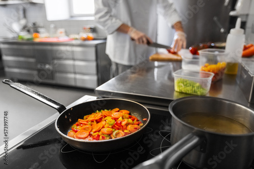 cooking food, profession and people concept - close up of frying pan with stewing vegetables and male chef at restaurant kitchen