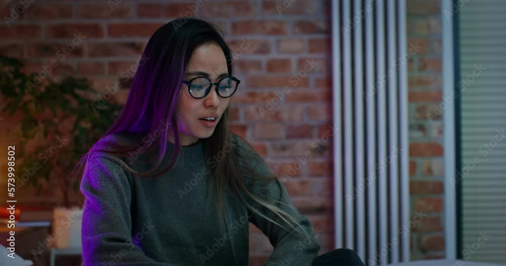 Young Asian woman wearing glasses smiling while video chatting on laptop, sitting on bed in stylish modern bedroom. The video moves from a long shot to a woman's face. Young Asian woman wearing glasses smiling while video chatting on laptop, sitting on bed in stylish modern bedroom. The video moves from a long shot to a woman's face.