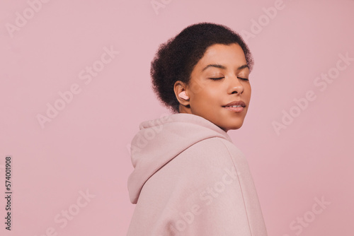 Stylish fashion african model in wireless earbuds standing with closed eyes on pink pastel background. Lifestyle concept in minimal style and copy space.