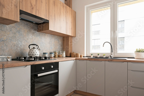 Interior of small kitchen with sink near window and wooden cabinets in kitchen furniture. Stove with gas and kettle. Modern pattern tiles on the wall.