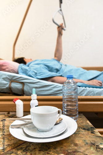 cup with saucer and bottle of water on table and sick person holding handle of homecare hospital bed on background at home photo