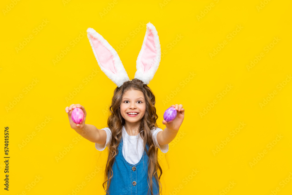 Fototapeta premium Happy Easter. A little girl holds two painted eggs for a spring holiday. A charming child with rabbit ears in a blue sundress on a yellow isolated background.