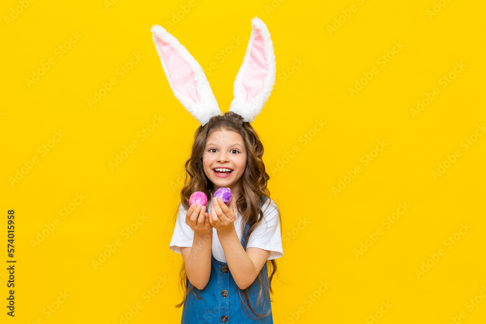 Happy Easter. A little girl holds two painted eggs for a spring holiday. A charming child with rabbit ears in a blue sundress on a yellow isolated background.
