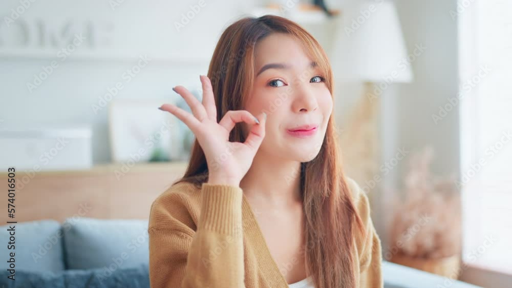 Attractive young asian woman smiles at camera showing hand OK sign in living room at home, Female shows everything fine success gesture body language, approval Attractive young asian woman smiles at camera showing hand OK sign in living room at home, Female shows everything fine success gesture body language, approval