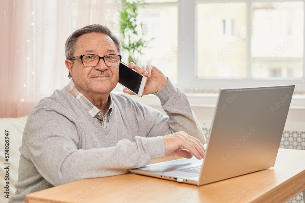 Cheerful senior man has phone conversation, chatting on the smartphone working with a laptop remotely from home. Cheerful senior man has phone conversation, chatting on the smartphone working with a laptop remotely from home.
