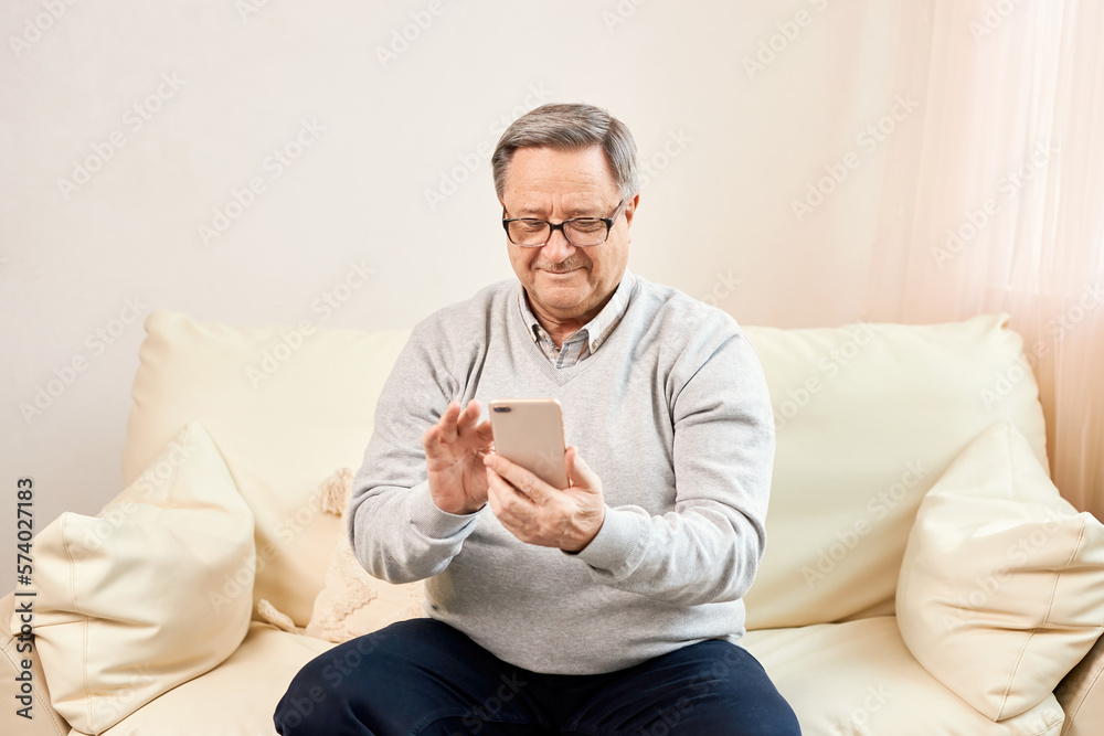 People And Technology. Portrait of smiling mature man using mobile phone, watching video or reading sms message, sitting on the couch in living room at home. Elderly man uses modern technology