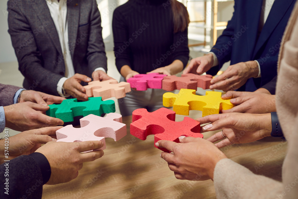 Cropped hands of a group of business people assembling big multicolor puzzle. Cooperation, teamwork, help and support concept. Coworkers join jigsaw pieces in the office above the wooden table. Cropped hands of a group of business people assembling big multicolor puzzle. Cooperation, teamwork, help and support concept. Coworkers join jigsaw pieces in the office above the wooden table.