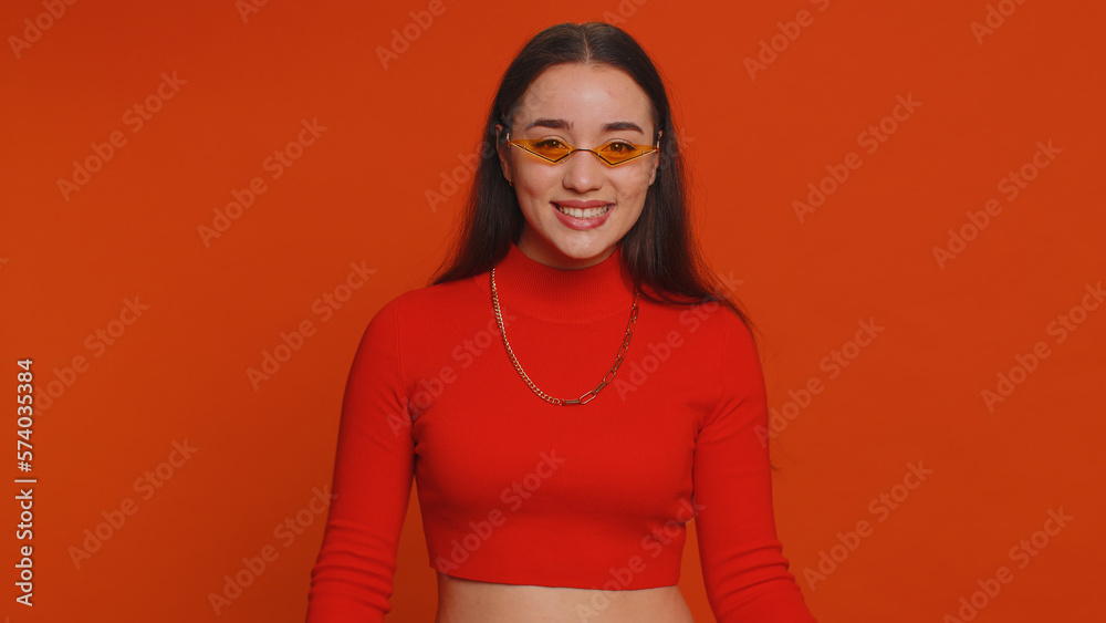 Fototapeta premium Portrait of happy pretty young woman in crop top wearing sunglasses, looking at camera with toothy charming smile, flirting, expressing optimism. Millennial girl isolated on red studio background