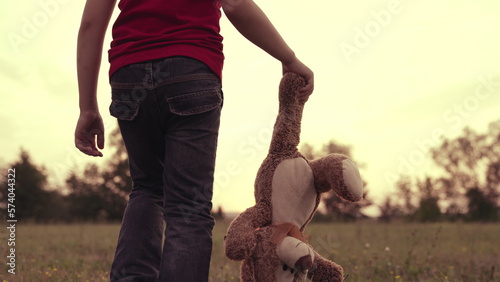 child carries teddy bear by leg sunset. offended orphan walks with doll park. boy son child alone with plush friend summer walk. boy walks lawn with teddy bear. childhood dream little child. no mood