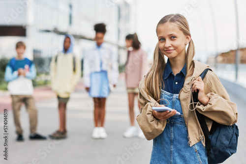 Blond pretty schoolgirl with backpack and smartphone looking at camera while standing against group of intercultural friends outdoor