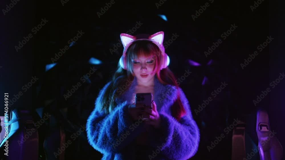 Cute gamer girl wearing headphones with luminous cat ears - using her phone in streaming studio Cute gamer girl wearing headphones with luminous cat ears - using her phone in streaming studio