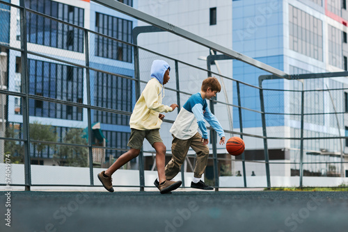 Two multicultural boys in activewear running after ball and trying to catch it while playing basketball at leisure or after school outdoors © Seventyfour