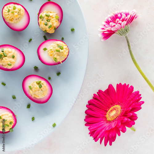 A platter of bright pink devilled eggs alongside two bright pink flowers.