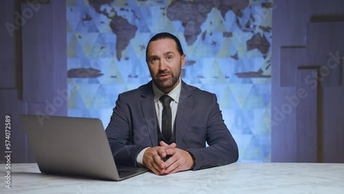 Stylish man presenter in suit and tie sitting at table, modern laptop, news, telling news to camera. In the background is a world map.