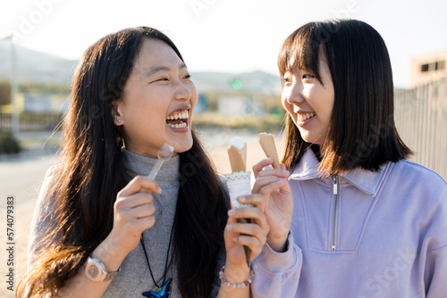 Happy lifestyle of a couple of young Asian women smiling at each other outside. The girls look at each other while eating an ice cream cone. Outdoor activity concept.