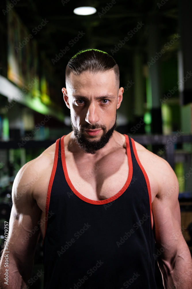 A handsome muscular male bodybuilder with a beard in a black T-shirt looks at the camera in the gym A handsome muscular male bodybuilder with a beard in a black T-shirt looks at the camera in the gym